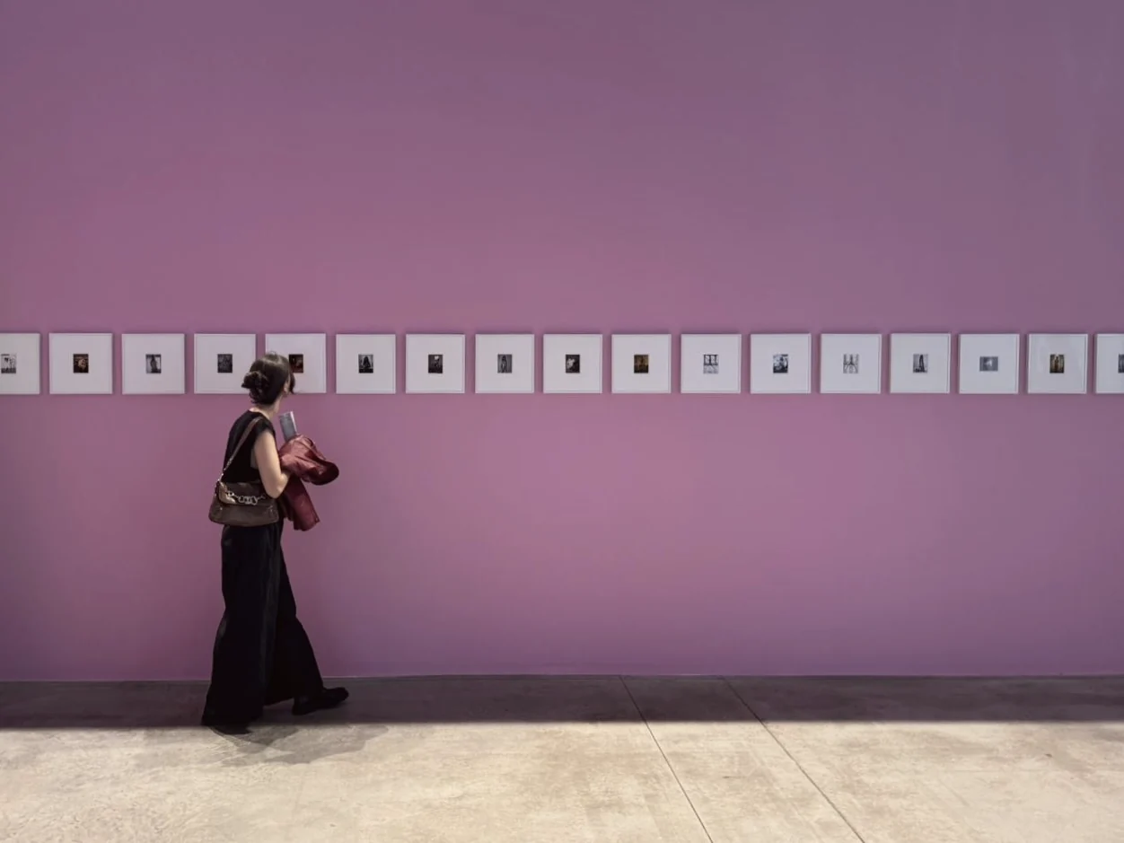 Ada walking past a row of small framed photographs on a pink gallery wall.